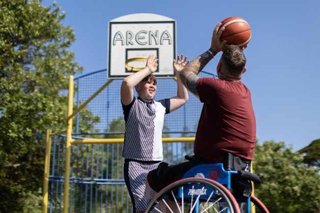 Aaron is sitting in his wheelchair on a basketball court with his son. His son is reaching for the basketball which defending the net. Aaron is throwing the ball.