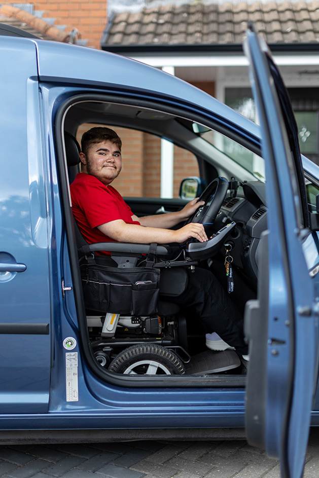 Harry is opening the front door of his vehicle smiling. Whilst he has his hands on the steering wheel. He is in his powered wheelchair.
