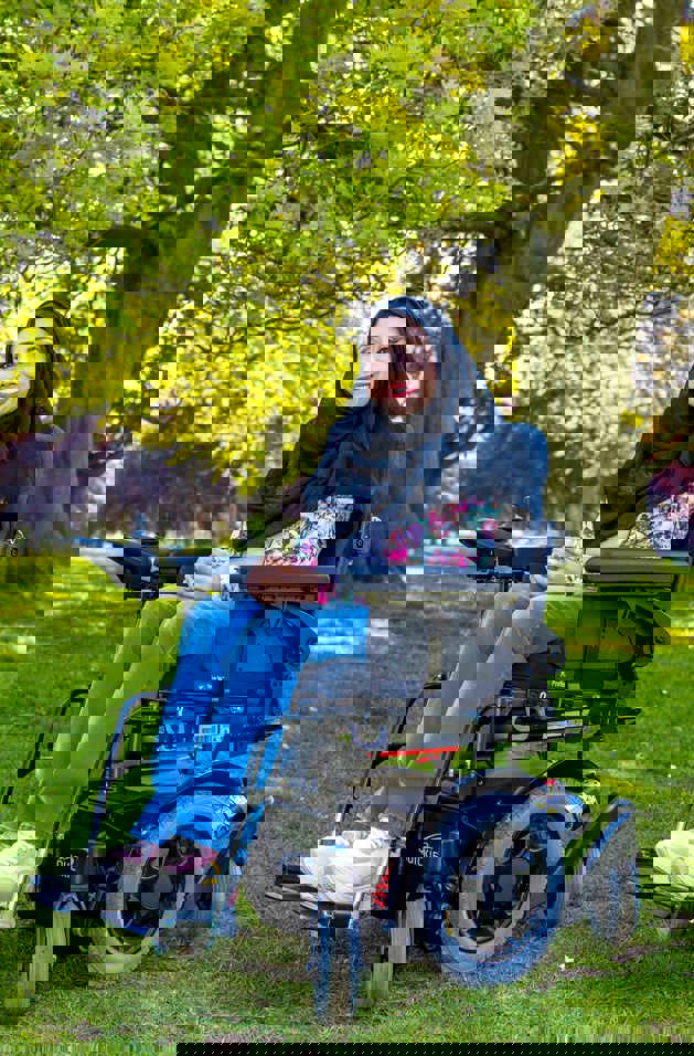 Nazmin is in her powerchair, outside on a sunny day. She's wearing a dark hijab, bright shirt and a big smile