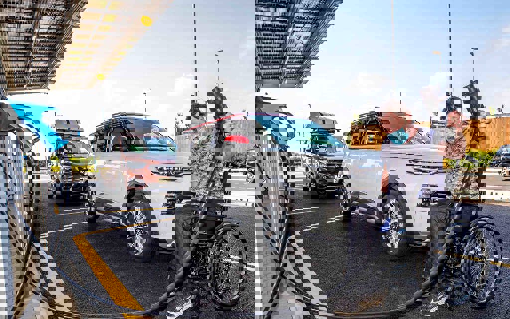 A man in a wheelchair is holding a charging plug for an electric car. The wheelchair is in front of a parked electric vehicle.
