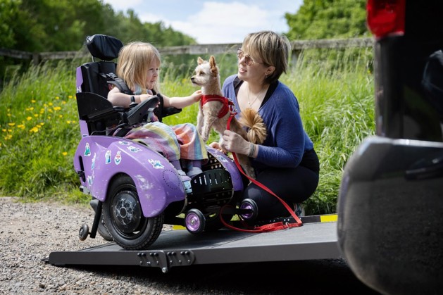 Poppy is sitting in her powered wheelchair on the ramp of her vehicle. Her mum Kim is crouched next to her, holding their family dog. Poppy is petting the dog and smiling.