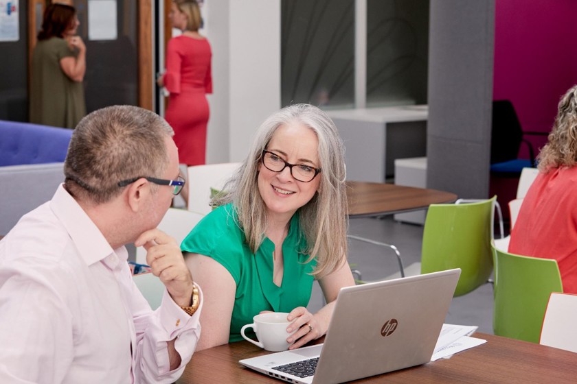 Two colleagues are meeting in a large office talking next to a laptop and printed papers.