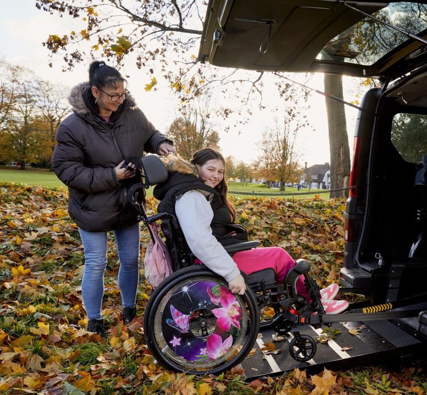 Becky is in her wheelchair on the ramp of her black wheelchair accessible vehicle. She is smiling to camera. She has long brown hair, black coat, grey hoodie, pink trousers and pink trainers. He mum Sarah is operating the ramp, she is has black long hair in a pony tail with a black coat, blue jeans and black trainers, she is smiling at Becky. Behind them is a park with orange and yellow leaves on the trees.
