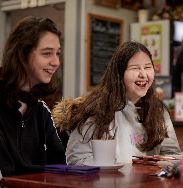 Holly and Becky are pictured sitting talking in a café, they each have a hot drink and are facing away from the camera smiling. Holly on the left hand side, she has long brown hair and is wearing a black hoodie. Becky is on the right hand side she has long brown hair and is wearing a grey hoodie.