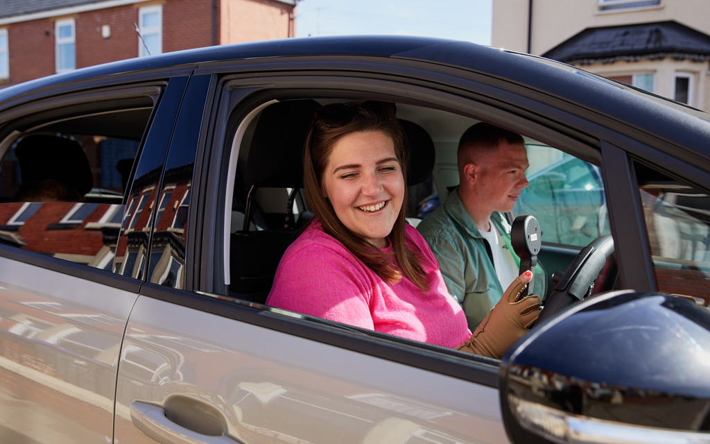 A woman sits in the driver's seat of a car holding a hand control while a man sits next to her as a passenger