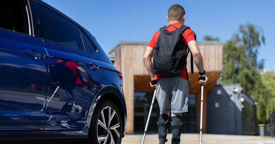 A young man is walking away from his blue vehicles using crutches. He had a red top on with a black backpack and blue shorts.