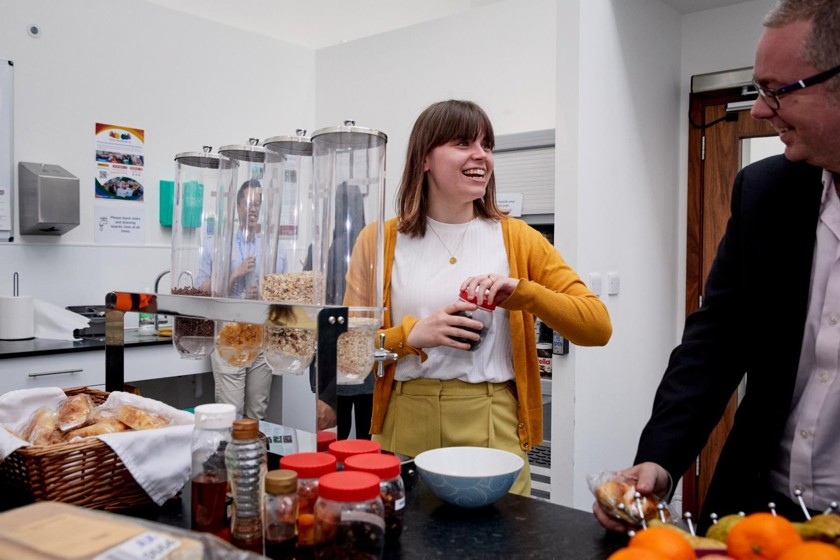 A lady is organising breakfast in an office. While talking and laughing with a colleague.