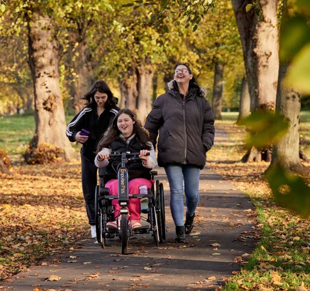 Becky, Holly and Sarah are in their local park, the leaves on the many trees around them are yellow and orange. Becky is wheeling in her wheelchair in the middle, whilst her sister Holly is to her left and her mum Sarah is to the right.