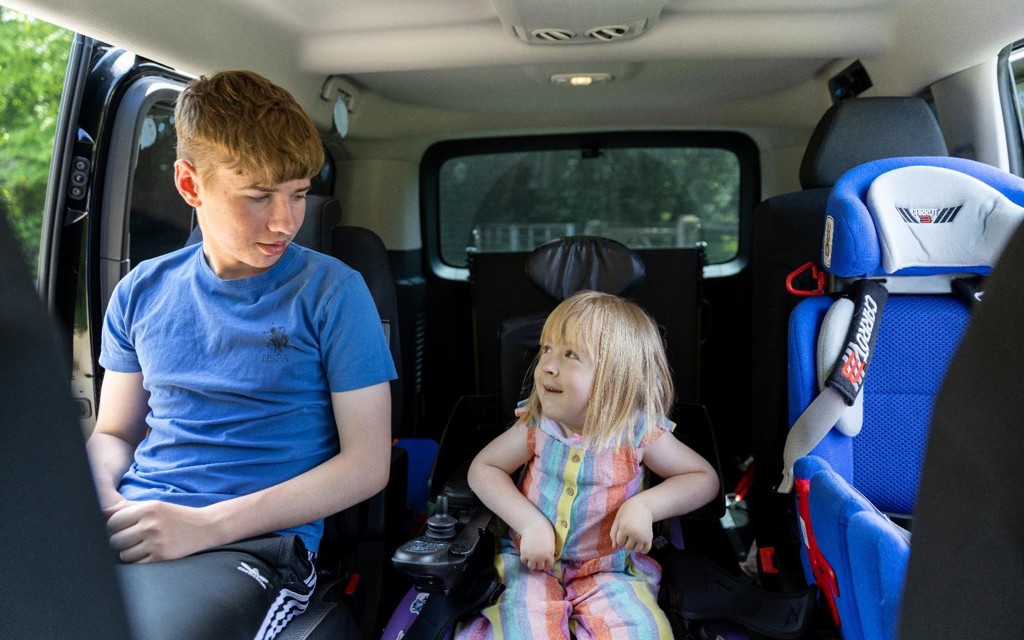 Poppy is sitting in her powered wheelchair in her vehicle. To the left of her is her old brother Liam, Poppy and Liam and smiling at one another. To the right of her is her car seat.