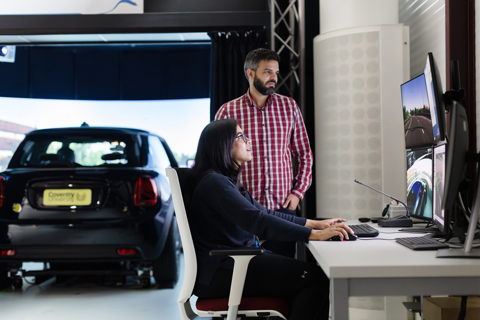 A woman and a man look at a computer screen with a car in the background