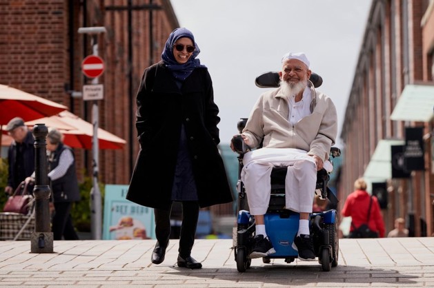 The image has red bricked buildings in the background with shops and big umbrellas from outside restaurant seating. Moosa's wife is on the left-hand side walking and talking with Moosa who is smiling on the right-hand side in his electric wheelchair.