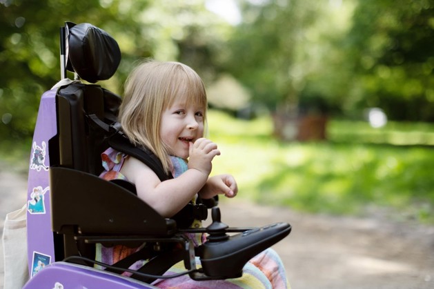 The image is a close up of Poppy in her lilac powered wheelchair. She is laughing. Behind her are lots of green trees.
